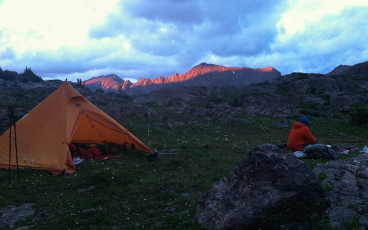 a person sits outside an orange tent while the sun rises, shining light on the mountains in the background